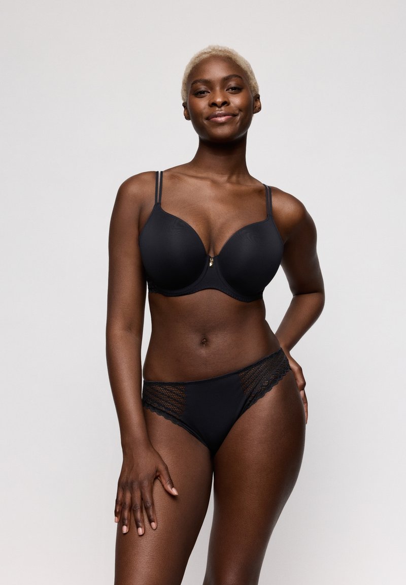 Zelfverzekerde vrouw met kort blond haar die een zwarte beha en kanten slipje met randjes showt tegen een effen lichte achtergrond.