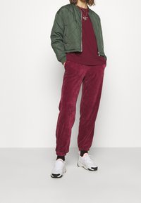 Veste bomber matelassée verte, t-shirt bordeaux et pantalon en velours côtelé bordeaux avec poignets élastiques. Baskets blanches avec des accents noirs et orange.