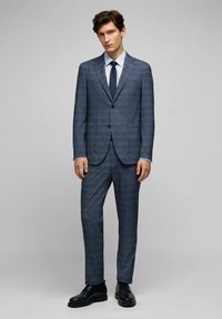 Marineblauw pak met een subtiel ruitpatroon, enkele knoopsluiting en rechte broekspijpen. Gecombineerd met een wit shirt en een donkere stropdas.