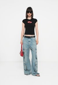 Black fitted t-shirt with a red logo, paired with light blue wide-leg cargo jeans and a red glossy handbag. Model wearing black sunglasses.