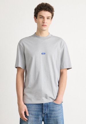 T-shirt en coton gris avec un col rond, des manches courtes et une étiquette de logo "HUGO" bleue sur la poitrine, assortie à un jean en denim.