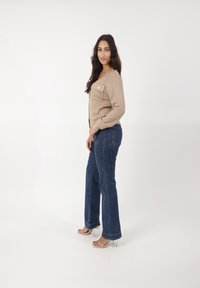 Beige textuur cardigan met een enkele voorzak, gecombineerd met donkerblauwe flared jeans en doorzichtige blokhaksandalen.