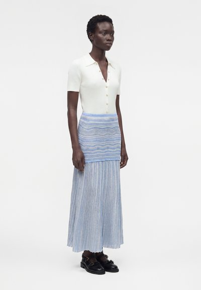 Γυναίκα με κοντά σγουρά μαλλιά που φοράει λευκό κοντομάνικο πουκάμισο με κουμπιά και μακριά μπλε φούστα με σχέδια και μαύρα παπούτσια.