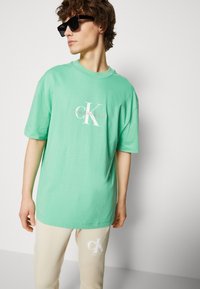 Camiseta de algodón verde menta con mangas cortas, presenta el logo blanco de "Calvin Klein Jeans" en el frente y en la parte posterior, combinada con pantalones beige pálido.