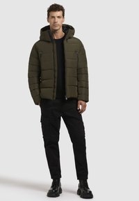 Geacă puffer de culoare verde măsliniu, cu quilt horizontal, glugă, buzunare cu fermoar, asociată cu pantaloni cargo negri și cizme cu șireturi.