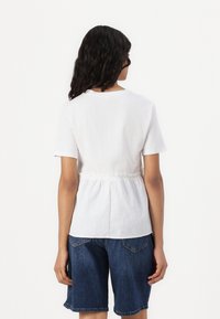 Hvit t-skjorte med korte ermer og samlet midje, kombinert med blå denimshorts. Stoffet ser glatt og lett ut.