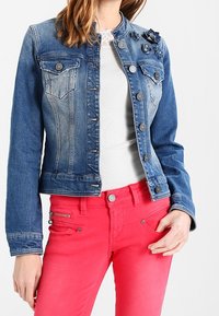 Džínová bunda s přechodem ze světle modré do tmavě modré, s knoflíkovým zapínáním, kapsami a květinovými doplňky na jednom rameni, nošená přes bílou košili.