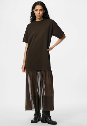 Brąsowa, oversize sukienka t-shirtowa z plisowanym, przezroczystym dołem. Górna część jest jednolita, natomiast dolna posiada przezroczystą teksturę. Czarne botki za kostkę.