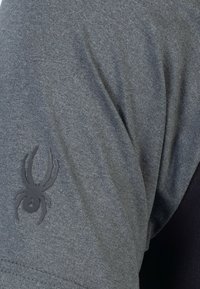 Camisa gris de manga corta con una textura suave, que presenta un logotipo de araña negra en la manga izquierda y un detalle de color contrastante en el lado.