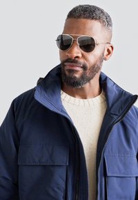 Giacca isolante blu navy con zip frontale, due tasche sul petto, indossata sopra un maglione lavorato a maglia color crema. Occhiali da sole argento con lenti scure.