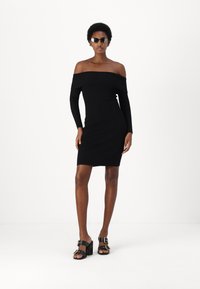 Abito nero a costine con scollo off-the-shoulder, design aderente e maniche lunghe. Abbinato a mule neri con tacco alto e dettagli in oro.