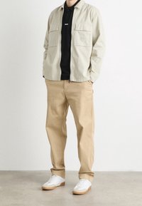 Beżowa kurtka koszulowa z dwoma przednimi kieszeniami, czarna koszulka polo pod spodem, beżowe spodnie i białe sneakersy z jasnobrązowymi podeszwami.