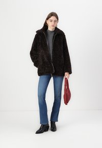 Veste en fausse fourrure marron avec un col haut, fermeture éclair sur le devant, associée à un haut gris et un jean bleu. Accessoirisée avec un sac à main rouge.