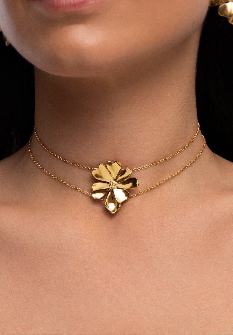 Collier ras-du-cou double chaîne en or avec un grand pendentif fleur poli porté autour du cou d’une femme sur un fond sombre.