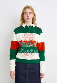 Zaļš, balts un oranžs svītrots polo krekls ar apkakli, ar "Miamas Universitātes" grafiku centrā un Adidas logotipu uz krūtīm.