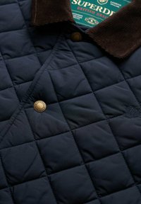 Chaqueta acolchada azul marino con cuello de pana marrón, que presenta un botón a presión dorado y un sutil detalle de logo bordado. Fabricado con un tejido cálido y texturizado.