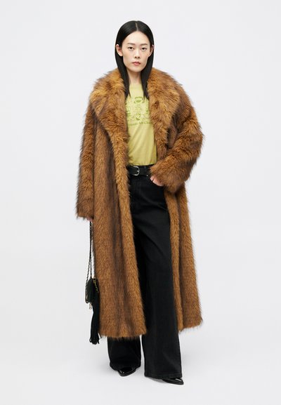 Žena v dlhom hnedom kabáte z umelej kožušiny, zelenom grafickom tričku, čiernych širokých nohaviciach a drží čiernu kabelku s reťazovým popruhom.