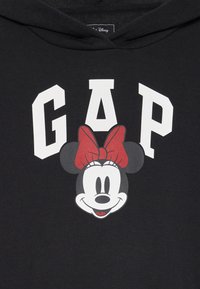 Crna dukserica s bijelim tekstom "GAP" i grafikom nasmijane Minnie Mouse s crvenom mašnom ispod teksta.