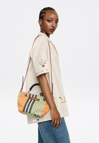 Colorful striped handbag with black and orange hues, featuring a curved handle, metal hardware, and a brand tag. Woven fabric texture visible.
