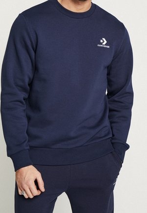Mężczyzna ubrany w granatową bluzę Converse i dopasowane spodnie, z małym białym logo Converse na lewej piersi.