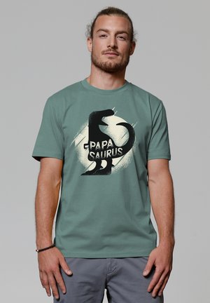 Groene katoenen t-shirt met een zwarte dino-silhouet en de tekst "PAPA SAURUS," met een lichte cirkelvormige spattenontwerpen erachter.