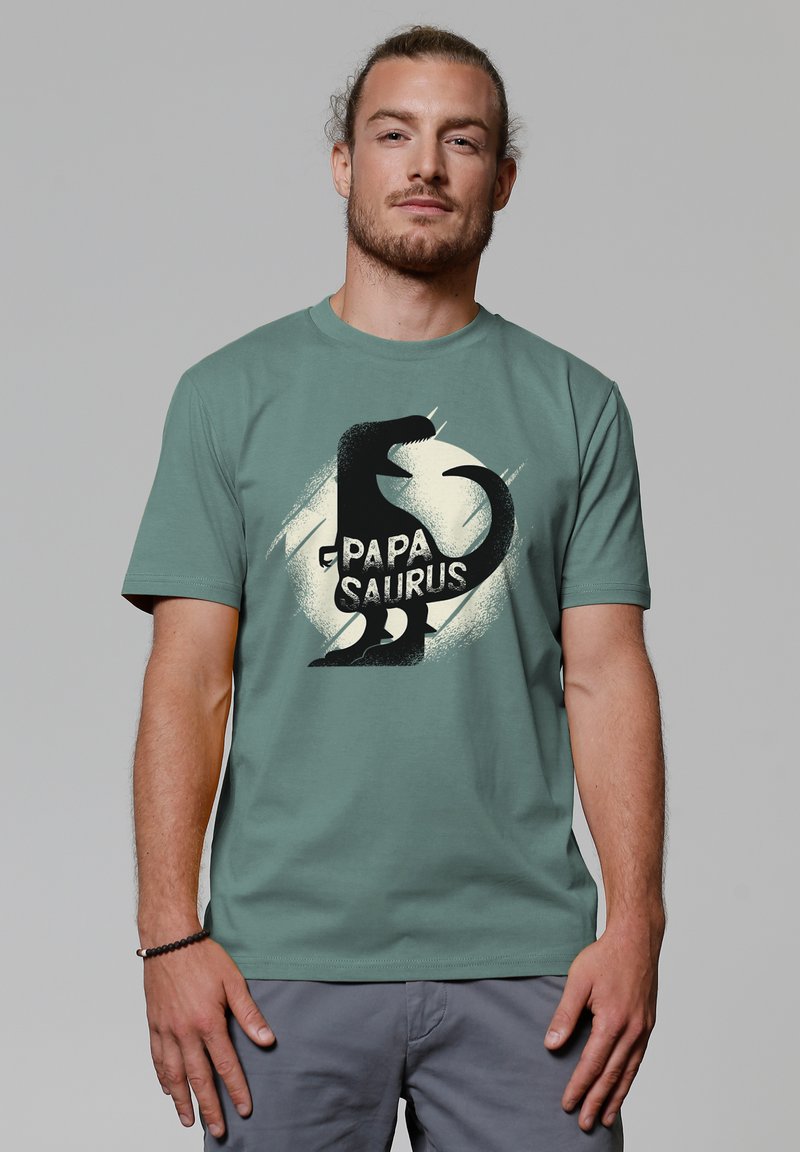 Groene katoenen t-shirt met een zwarte dino-silhouet en de tekst "PAPA SAURUS," met een lichte cirkelvormige spattenontwerpen erachter.