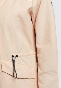 Light peach jacket with a smooth texture, featuring a front pocket, adjustable drawcord, and black hardware accents.