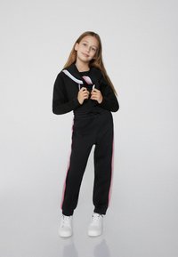 Schwarzer Sweatshirt mit koloriertem Kragen in Weiß und Pink sowie passenden Jogginghosen mit seitlichen Streifen. Abgerundet mit weißen Sneakers.