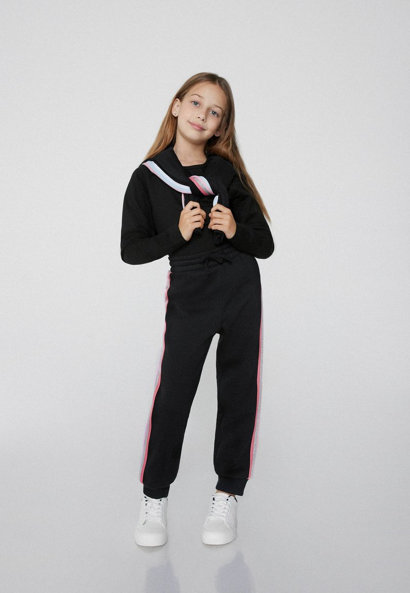 Schwarzer Sweatshirt mit koloriertem Kragen in Weiß und Pink sowie passenden Jogginghosen mit seitlichen Streifen. Abgerundet mit weißen Sneakers.