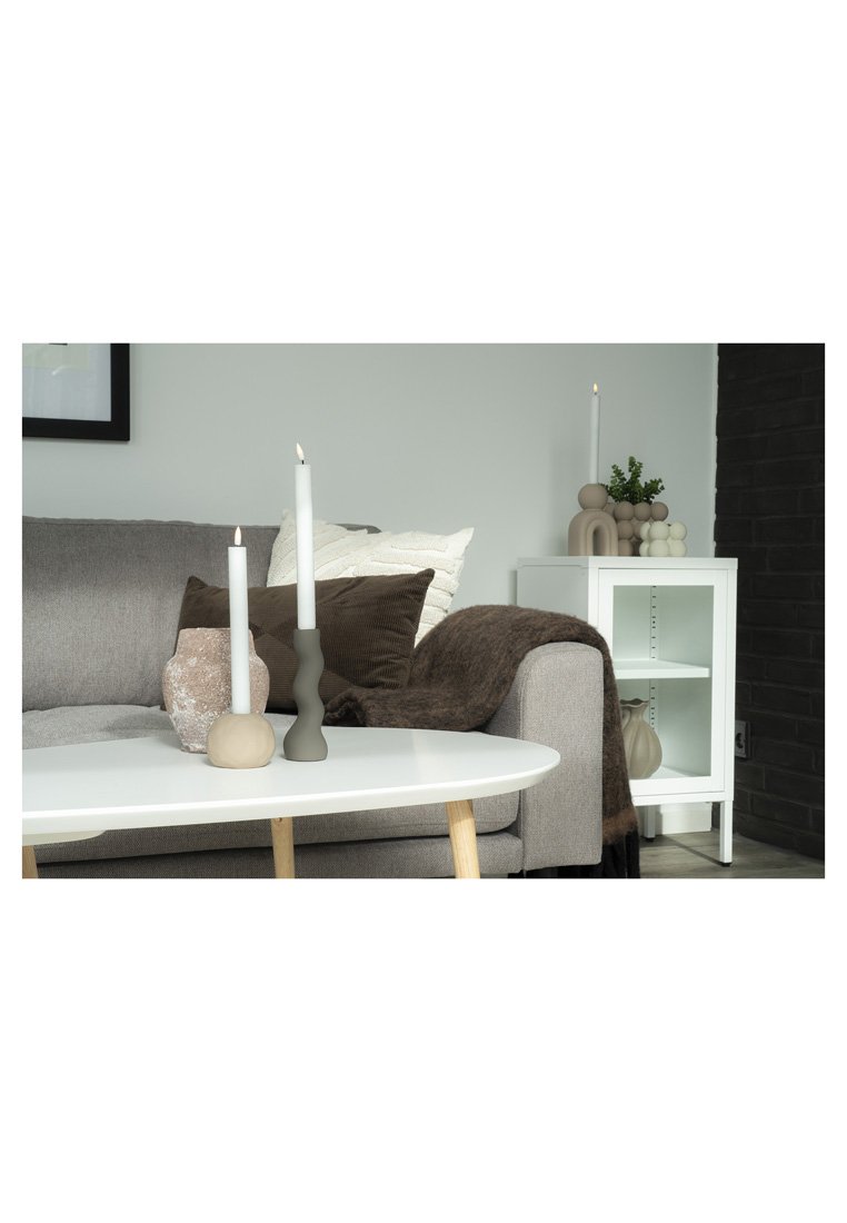 Divano grigio con cuscini marroni e bianchi, candele accese su un tavolino bianco, mobiletto bianco con vasi decorativi e pianta verde.