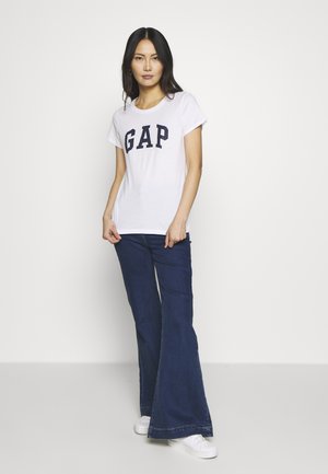 Bijela majica s mornarsko plavim "GAP" logotipom, u kombinaciji s tamnim trapericama širokog kroja. Model nosi bijele tenisice i stoji naslonjen na jednostavnu pozadinu.