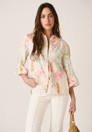 Mujer con blusa de bordado floral en crema y pantalones blancos, sosteniendo un bolso marrón claro sobre un fondo neutro.