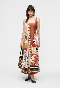 Γυναίκα που φοράει αμάνικο φόρεμα με τετράγωνη λαιμόκοψη και πολύχρωμα αφηρημένα σχέδια, κρατώντας μια καρό τσάντα και φορώντας μαύρα φλατ παπούτσια.