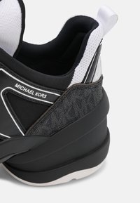 Talons de baskets Michael Kors noir et blanc avec tissu gris foncé texturé, semelle noire épaisse et détail en maille blanche au niveau du col de la cheville.