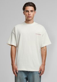 Tânăr purtând un tricou alb lejer cu textul „Slow Sunday Club” pe piept și blugi albastru deschis, stând în fața unui fundal gri.