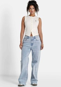 Top bianco senza maniche con chiusura a bottoni e dettaglio del logo, abbinato a jeans a gamba larga blu chiaro e sneakers grigie, su uno sfondo neutro.
