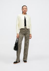 Cardigan crème avec boutons, haut noir, pantalon marron et beige à motifs, sac noir et chaussures noires à semelle marron.