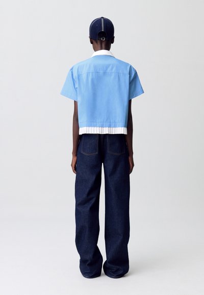 Camisa corta de manga corta en azul claro con dobladillo plisado blanco, combinada con jeans de pierna ancha en azul oscuro. Costuras visibles y detalle en el cuello trasero.