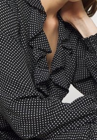 Svart blus med vita prickar. Har rynkad halsringning och ärmslut, mjukt tyg och avslappnad passform. Mönster betonar design detaljer.