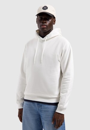 Jonge man die een witte hoodie en lichtblauwe spijkerbroek draagt, met een witte pet voorzien van een zwart logo, staand tegen een effen achtergrond.