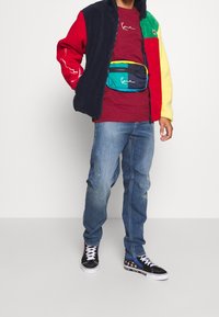 Fleecová bunda v námořnické modři, červené a žluté barvě se zipem, doplněná červeným tričkem. Modré džíny a černé tenisky s barevnými detaily dokončují outfit.