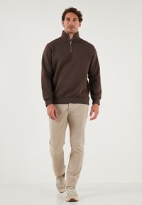 Bruine zip-neck trui van zacht materiaal, met lange mouwen en geribbelde manchetten. Gekoeld met lichtbeige broeken en beige sneakers.