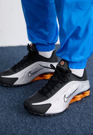 Osoba nosząca czarno-białe sneakersy Nike Shox z pomarańczowymi amortyzatorami i niebieskie spodnie stoi na niebieskim dywanie.