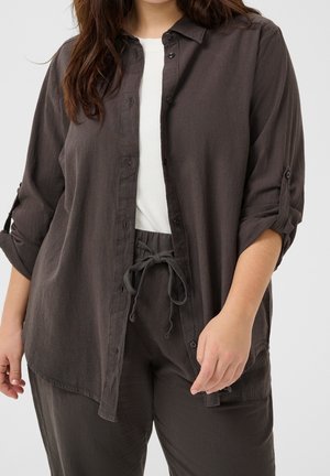 Donna che indossa una camicia grigio scuro con bottoni e maniche arrotolate, pantaloni coordinati con coulisse e una maglietta bianca sotto.