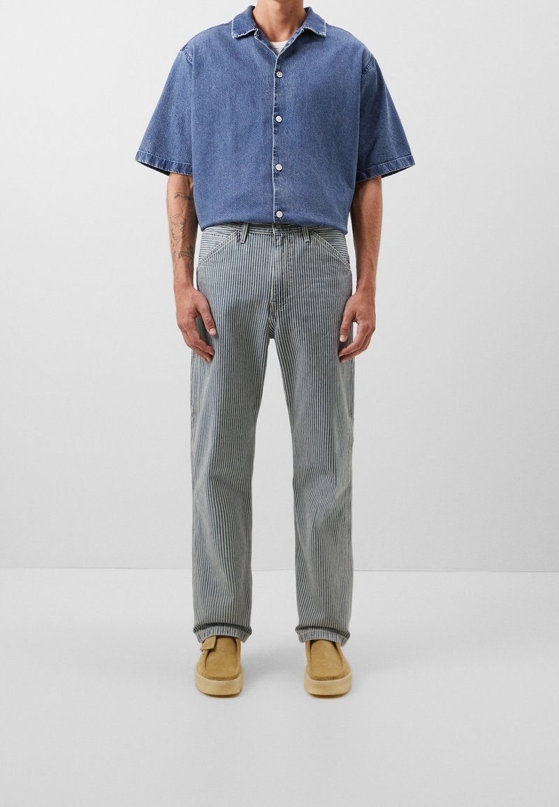 Hombre con camisa de mezclilla azul de manga corta, pantalones a rayas gris y blanco, y zapatos de ante color marrón, de pie frente a un fondo gris liso.
