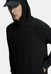 Musta huppari, jossa on sileä pinta ja raglan-hihat. Siinä on kenguru-tasku ja minimaalista ompelu-urheilua.