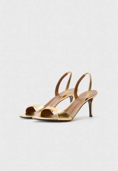 Złote metaliczne szpilki typu slingback z otwartym palcem, smukłym obcasem i miękkim wyściółką. Posiadają krzyżujące się paski i gładką teksturę.