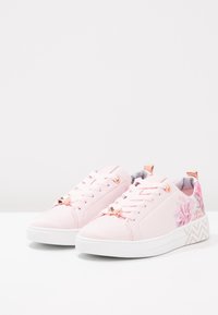 Baskets synthétiques roses avec accents à imprimé floral, œillets métalliques, et semelle blanche texturée avec un motif en zigzag. Lacets plats et col rembourré.