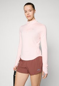 Lys rosa langermet treningsgenser med halv glidelås og reflekterende logo, kombinert med brune shorts med blå aksentlogo.