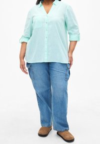 Světlá mátově zelená košile na knoflíky s výstřihem do V a kapsou na prsou; v kombinaci s volnými modrými džínovými cargo kalhotami a hnědými nazouvacími botami.